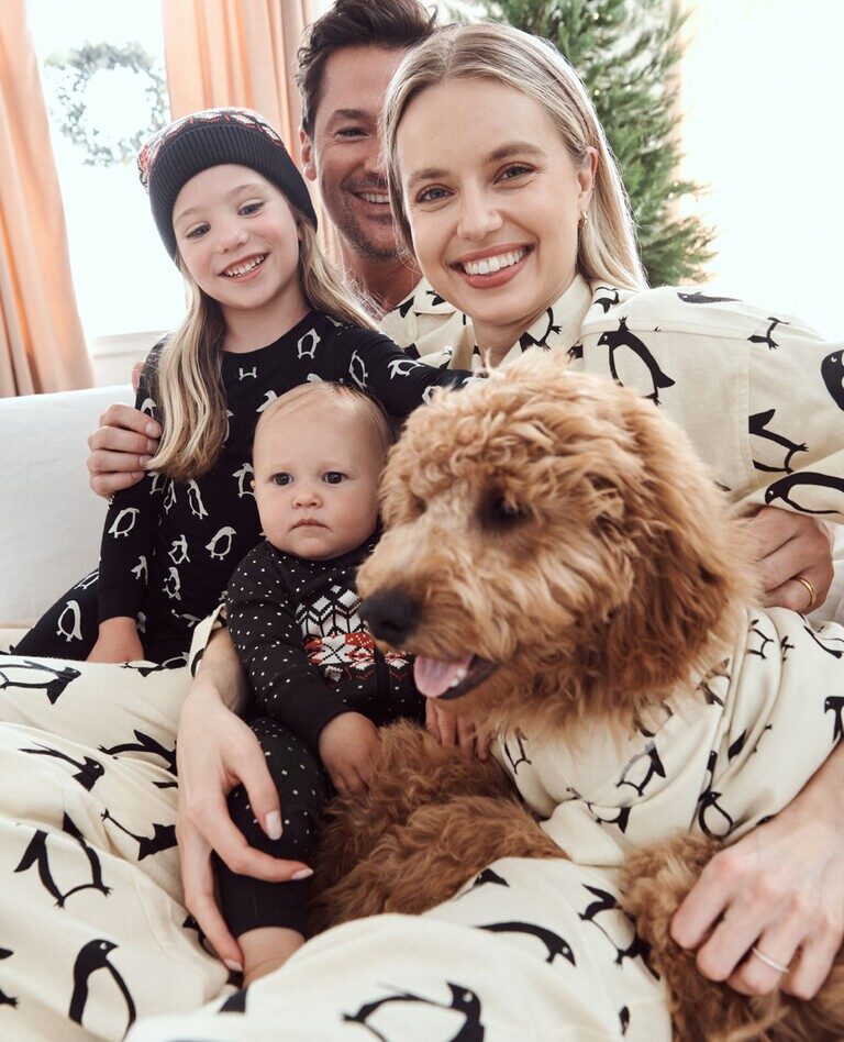 A family of four, including a mother, father, daughter, and son, as well as a family dog, sit on a white couch with matching penguin-themed pajamas. 