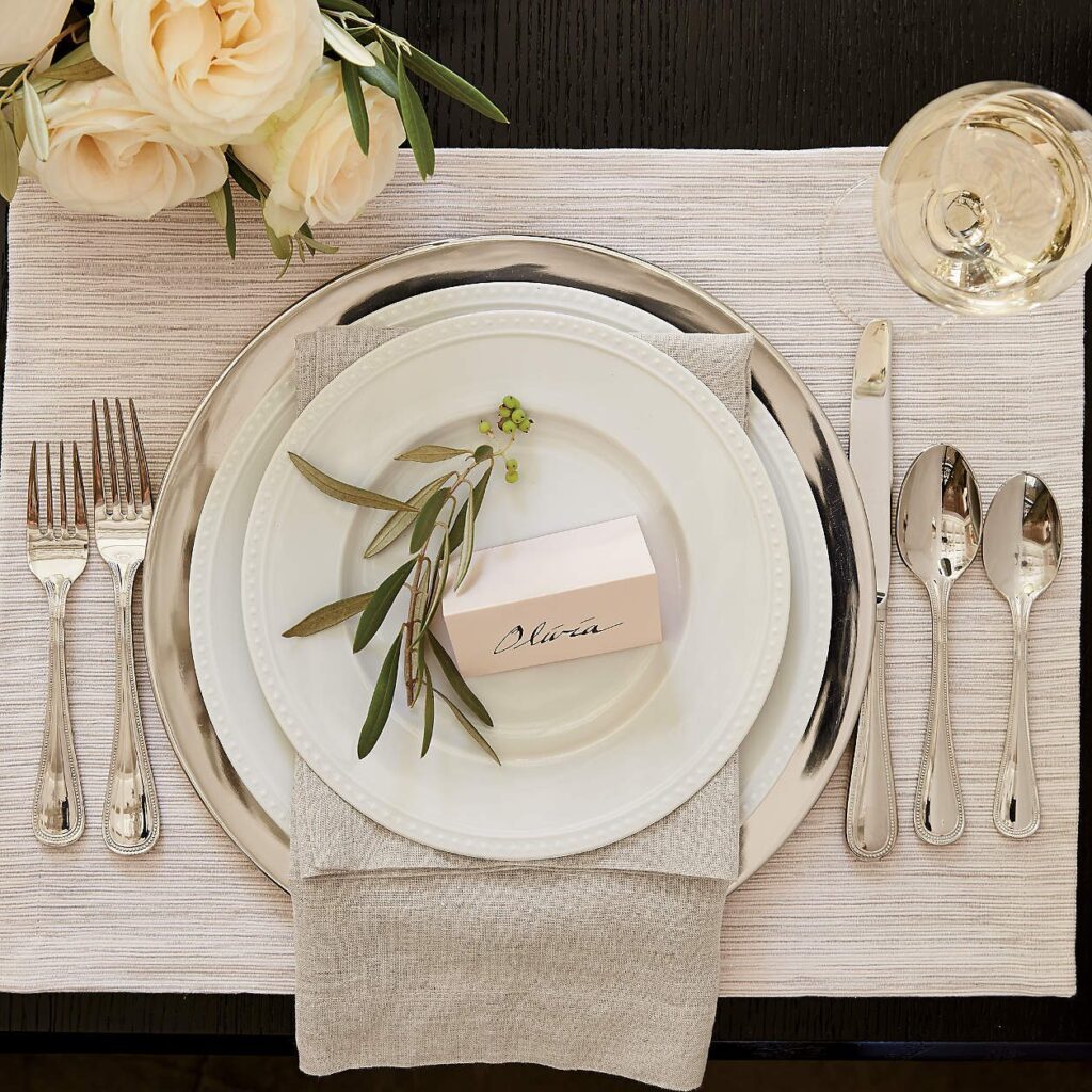 An elegant place setting on a table with a stack of plates, various silverware, white flowers, and a glass of wine.