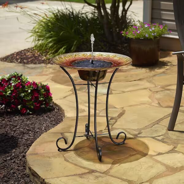 A glass solar fountain birdbath with a sunflower design shoots a jet of water while standing on a garden patio in full sunlight.